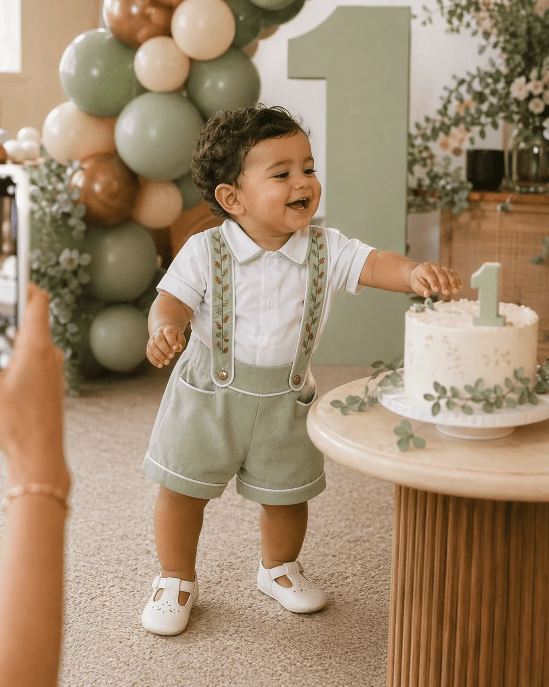 Bebê rindo ao lado do bolo de aniversário de 1 ano · Conjunto Versátil verde sage com suspensório bordado e sapato social branco · balões sage, terracota e caramelo · ocasião de mesversário